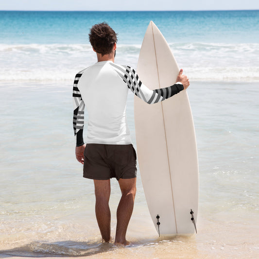 young guy looking at the beach and holding a white surfboard and wearing a white long sleeve rash guard with black and grey designs on the sleeves.