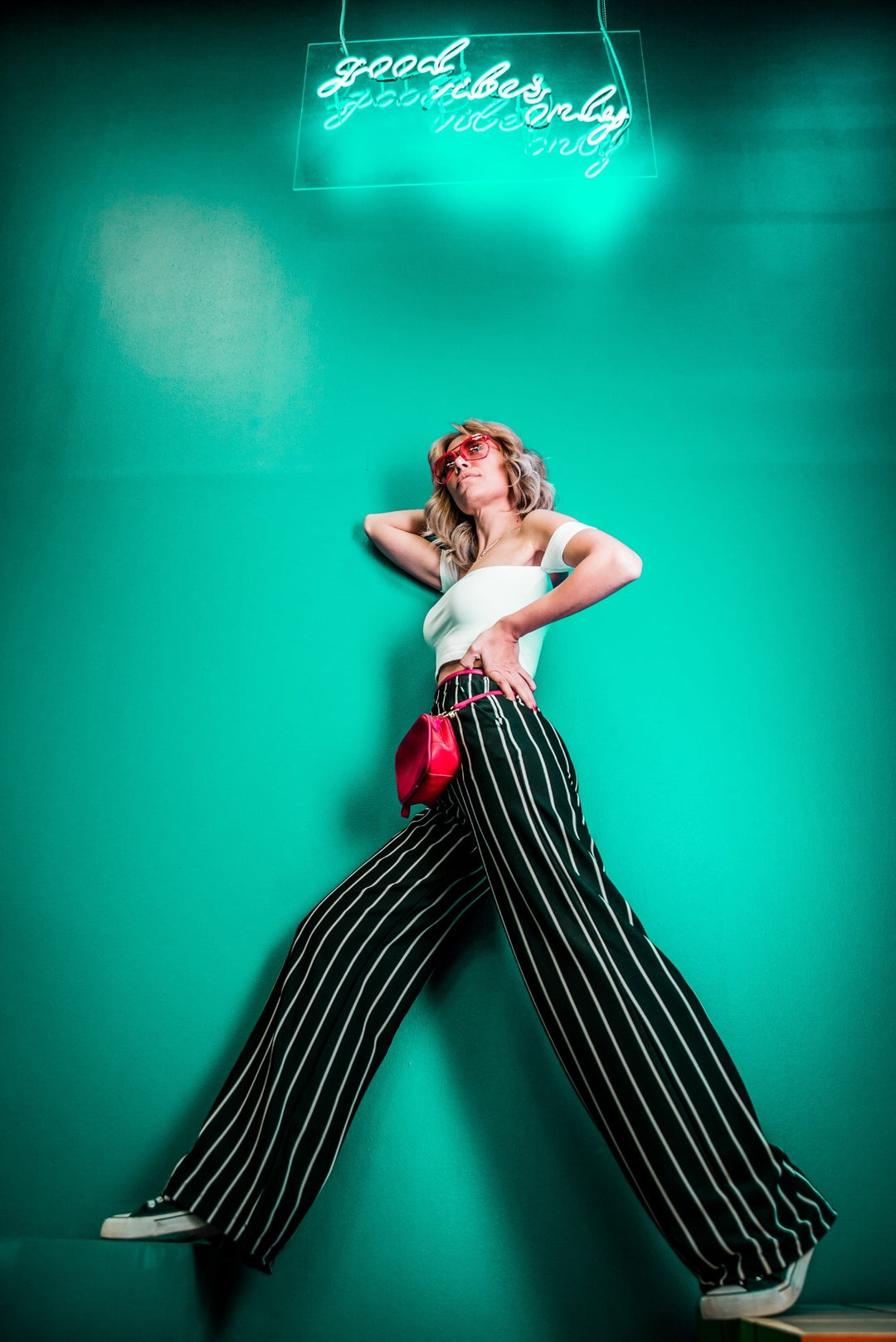 chica con una camisa de manguillo blanca y pantalones negros con lineas blancas posando en contra fondo con una pared aqua 